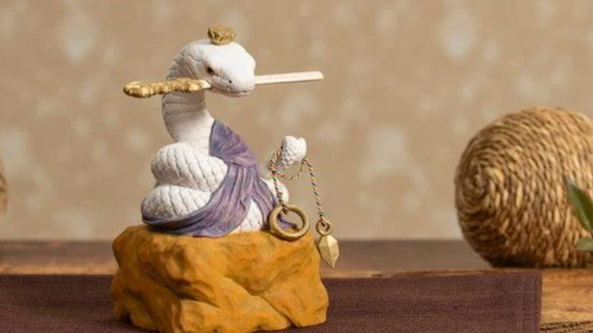 Decorative white snake figurine wearing a purple cloth, displayed on a golden base beside a woven ball on a tabletop.