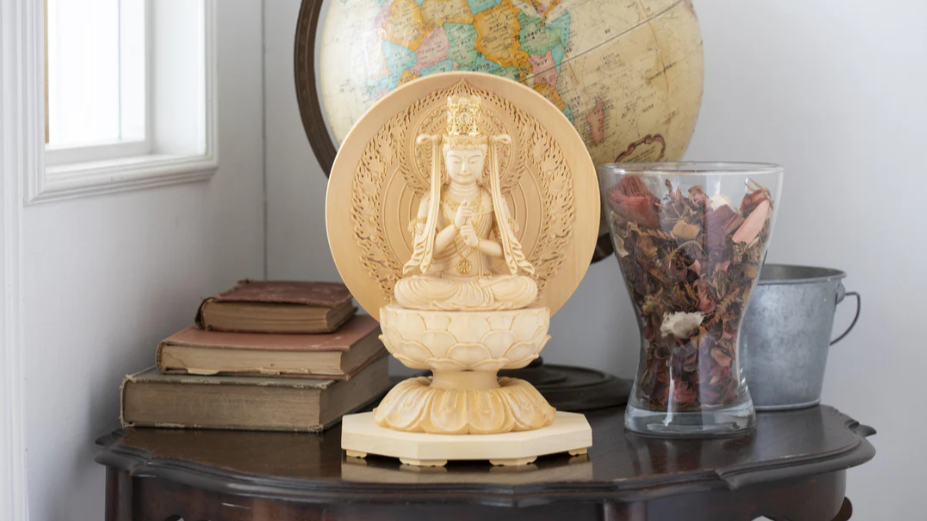 Ivory Buddhist statue displayed on a dark wooden table beside stacked books, a glass container of dried flowers, a metal bucket, and a vintage globe.
