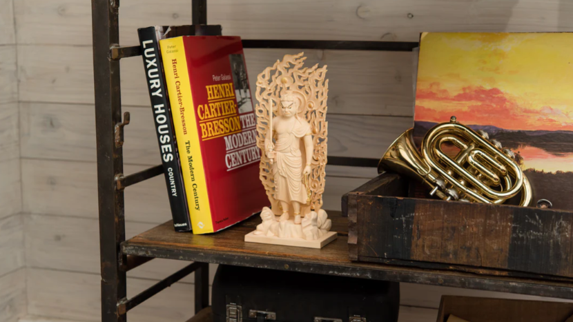 Small wooden Buddhist statue displayed on an industrial shelf beside books, a brass horn in a wooden box, and a sunset painting.