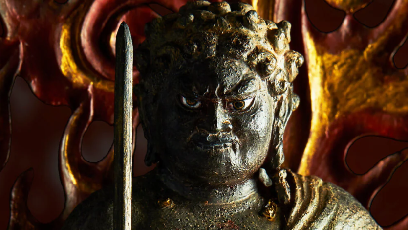 Close-up of a fierce Buddhist guardian statue holding a sword, with a red and gold flame halo in the background.