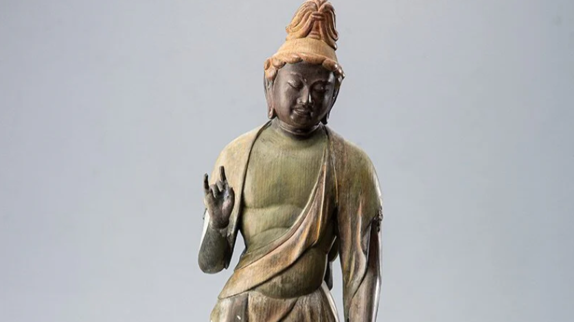 A weathered Buddhist statue with one hand raised, shown against a plain light gray background.