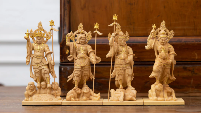 Four small wooden Buddhist guardian statues displayed in a row, each standing on an animal base and holding a staff, with a wooden cabinet in the background.