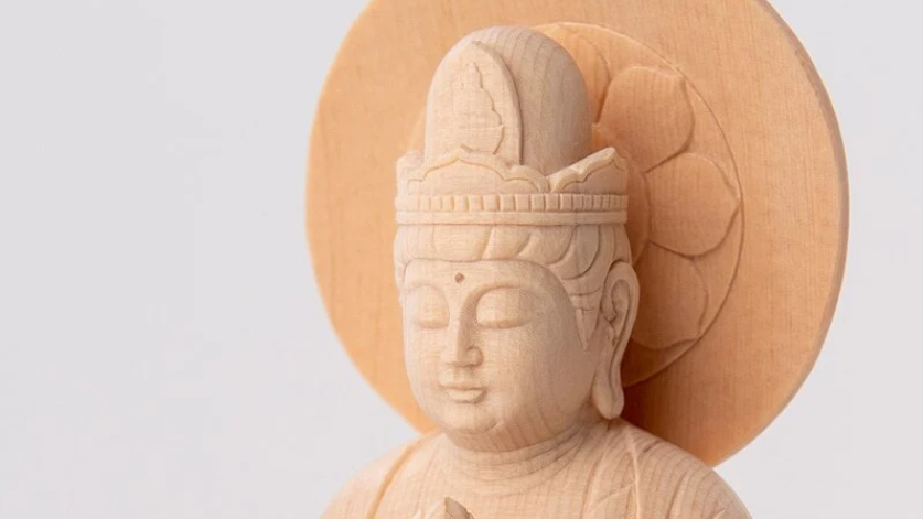Close-up of a light wooden Buddhist statue with a serene face and round halo against a plain white background.