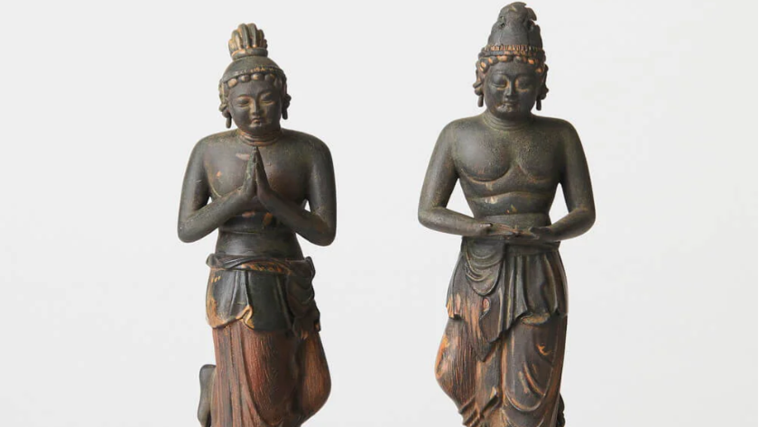 Two weathered Buddhist statues standing side by side against a plain white background, one with hands pressed together and the other holding hands at the waist.