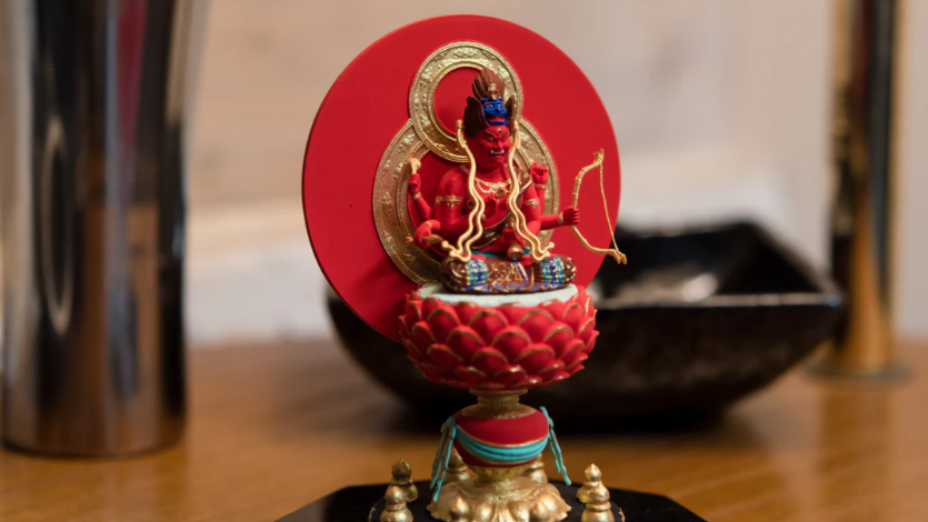 A small red Buddhist statue seated on a lotus pedestal, holding a bow with a red circular halo behind it.