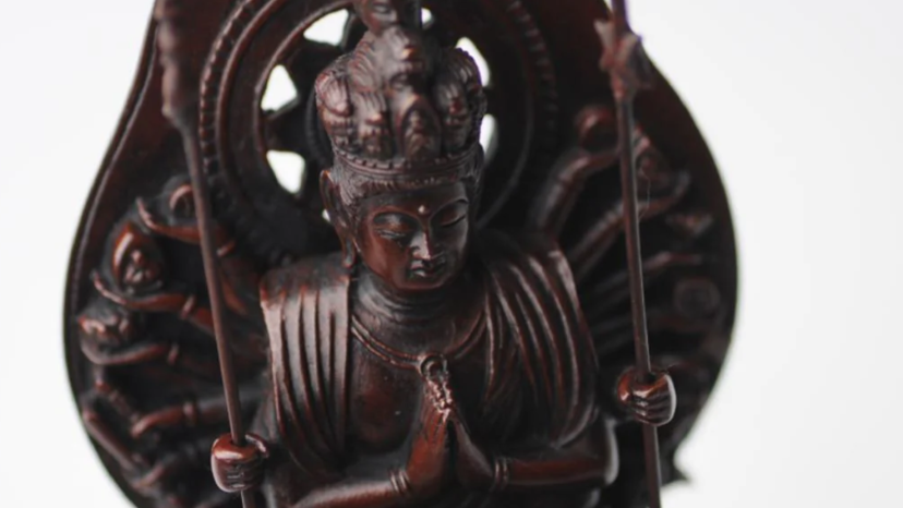 A dark brown Buddhist statue with hands in prayer, holding staff-like objects, with an openwork halo behind it.