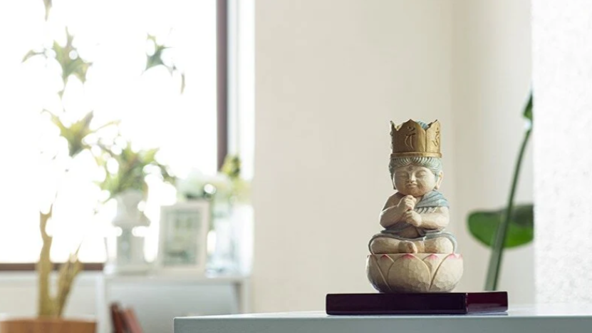 A small Buddhist statue with hands in prayer, placed on a shelf in a bright room.
