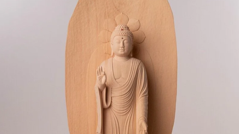 A pale wooden Buddha statue standing with one hand raised, set against a wooden back panel.