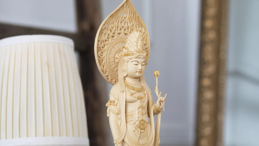 A pale wooden bodhisattva statue standing with a lotus flower and an ornate halo behind it.