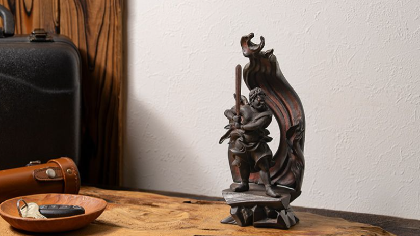 A dark Fudo Myoo statue standing with a staff, framed by a flame-shaped halo on a wooden desk.