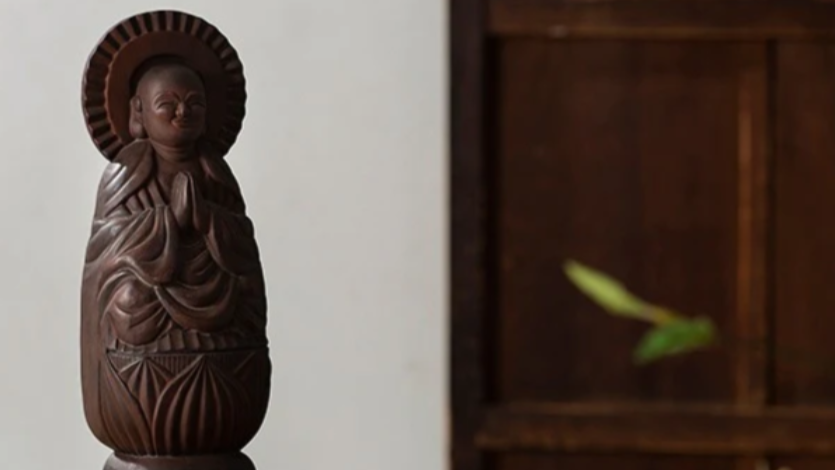 A small carved wooden Buddhist statue with hands in prayer, set against a dark wooden shelf with a round halo behind it.