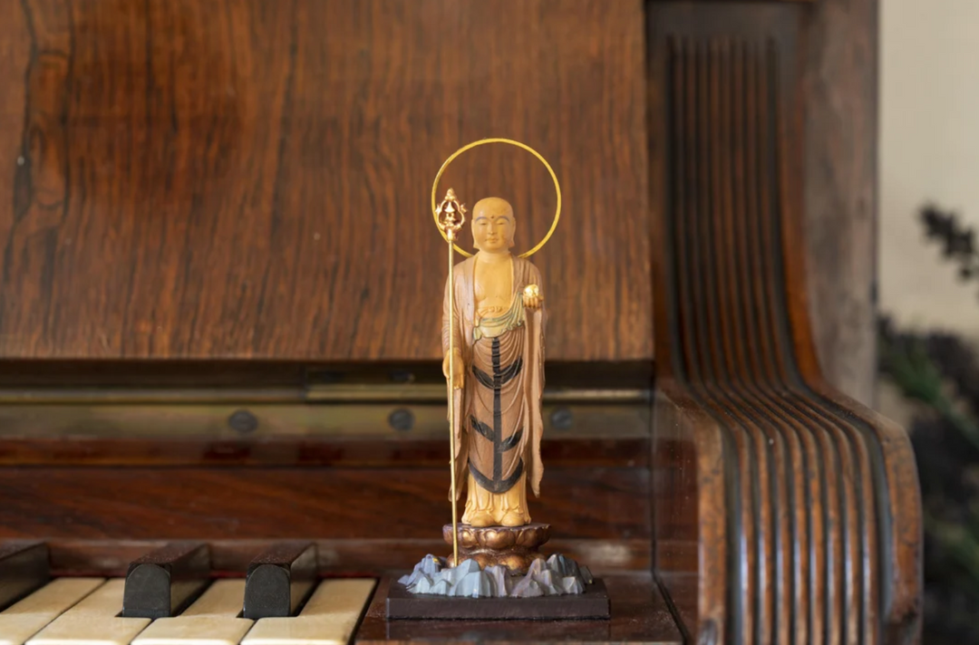 Standing Jizo statue with a halo and staff, displayed in a quiet interior setting, representing compassion, protection, and devotional offerings in Japanese Buddhism.