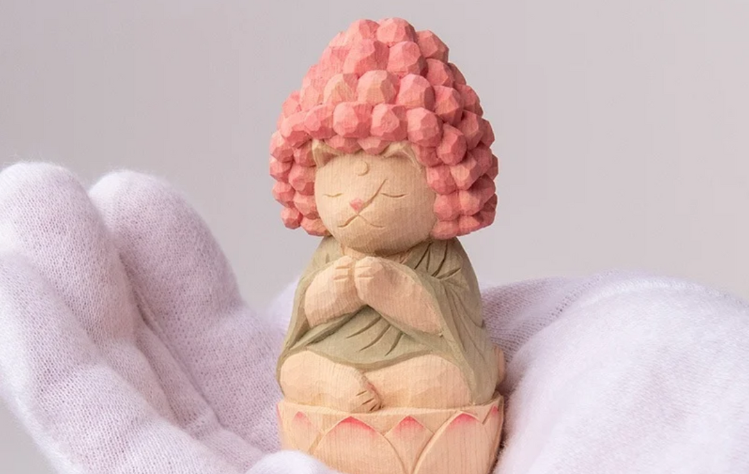 Whimsical wooden Buddha-inspired cat figurine with pink curled hair, seated on a lotus base, evoking traditional Buddha statue iconography.