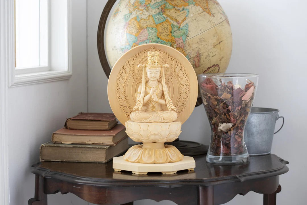 Wooden seated Kannon Bodhisattva statue on a lotus pedestal displayed on a wooden table with books, a globe, and dried flowers