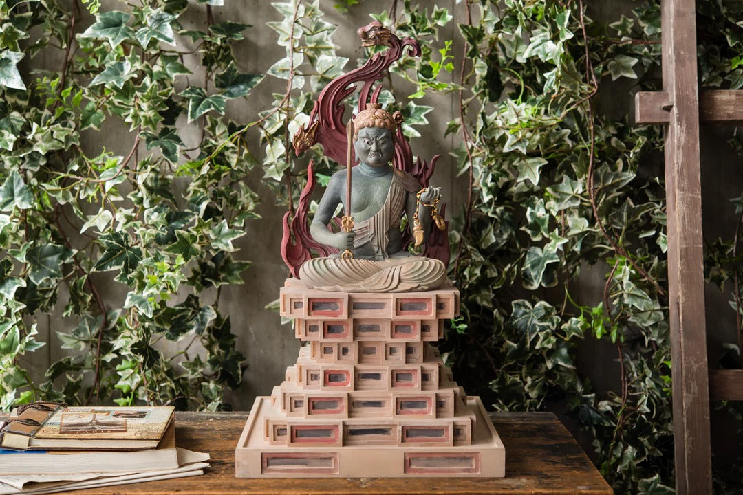 Wooden Wisdom King (Myo-o) statue seated on a pedestal with a flaming halo, displayed against a natural backdrop
